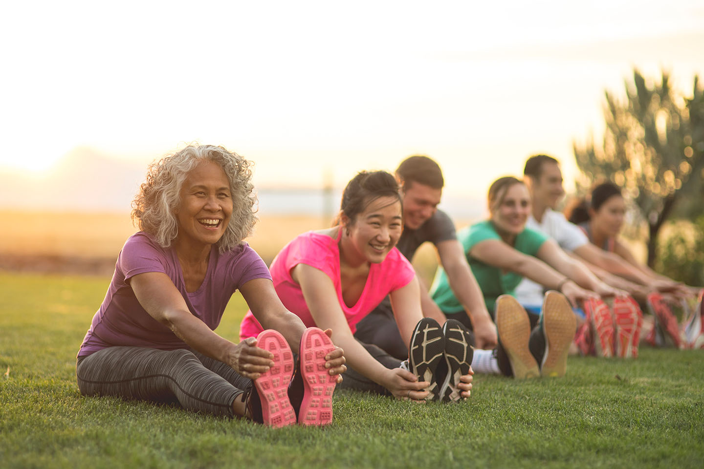 A group of adults attending a fitness outdoors are doing leg stretches. The participants are arranged in a line. The focus is on a mature woman who is smiling toward the camera.